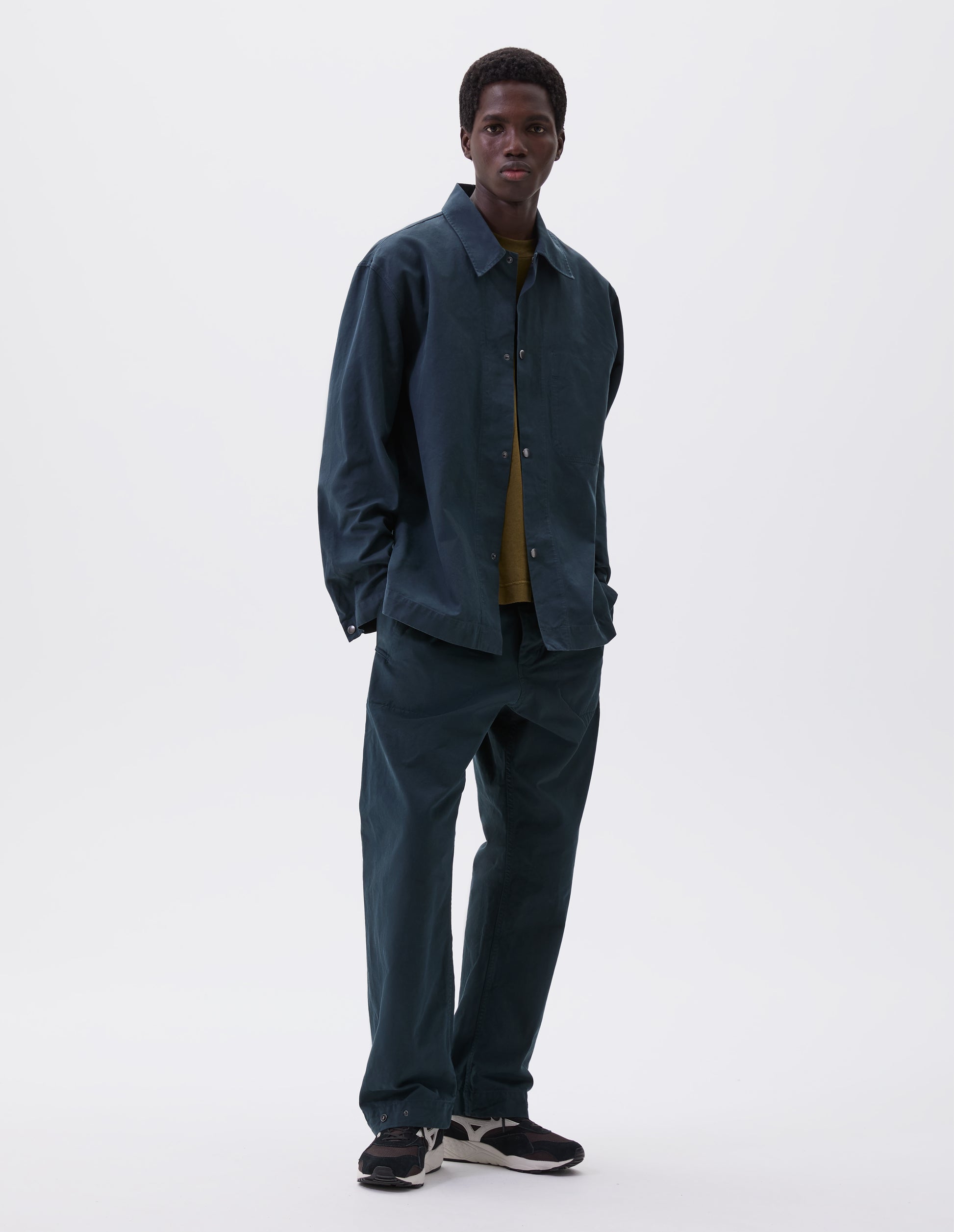 Full body image, front view of model wearing teal blue overshirt with press stud fastenings, stitch through chest pocket and adjustable cuffs. Styled with matching trousers, green t-shirt and brown and black trainers.