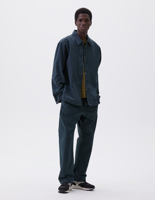Full body image, front view of model wearing teal blue overshirt with press stud fastenings, stitch through chest pocket and adjustable cuffs. Styled with matching trousers, green t-shirt and brown and black trainers.