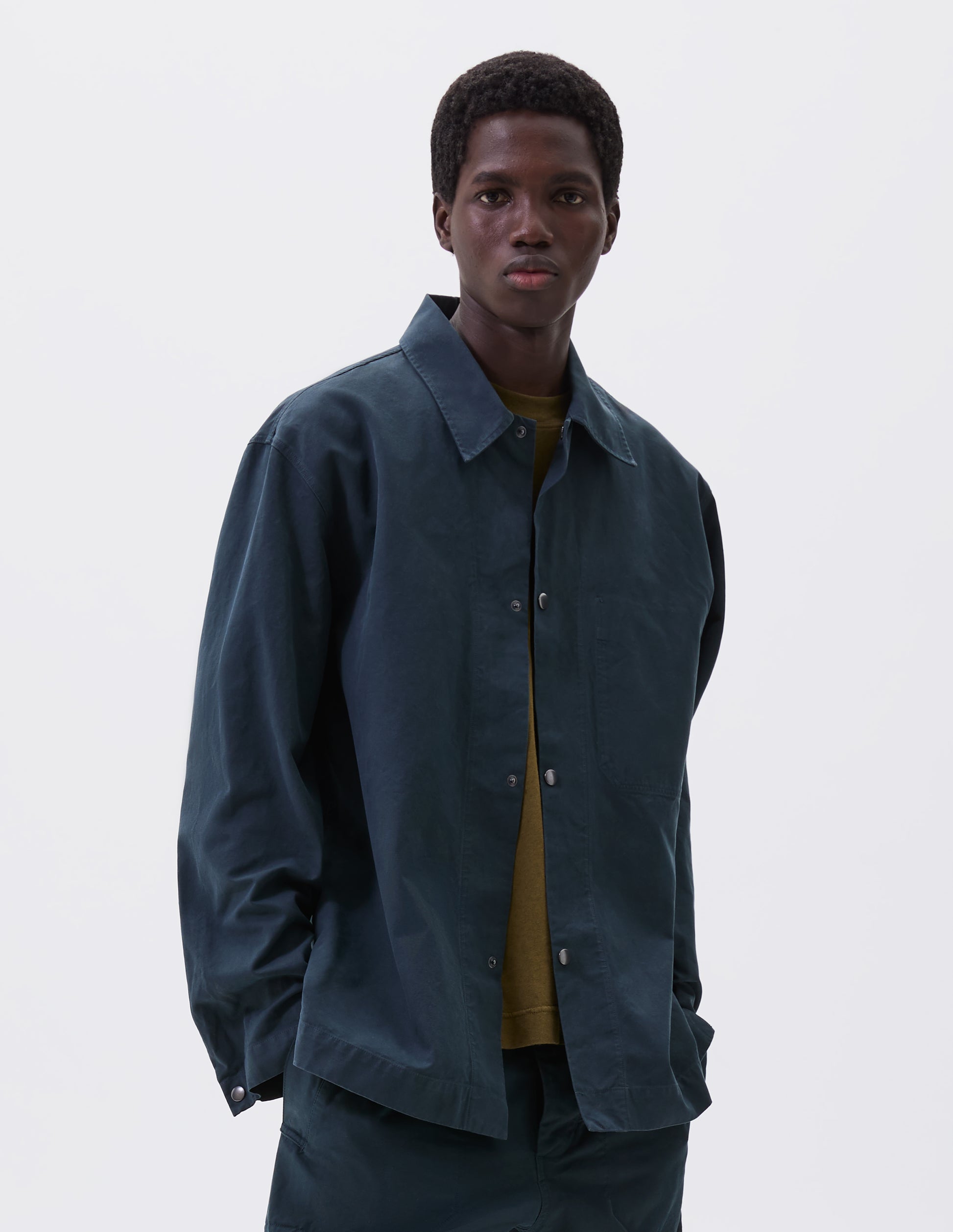 Cropped body image, front view of model wearing teal blue overshirt with press stud fastenings, stitch through chest pocket and adjustable cuffs. Styled with matching trousers and green t-shirt.