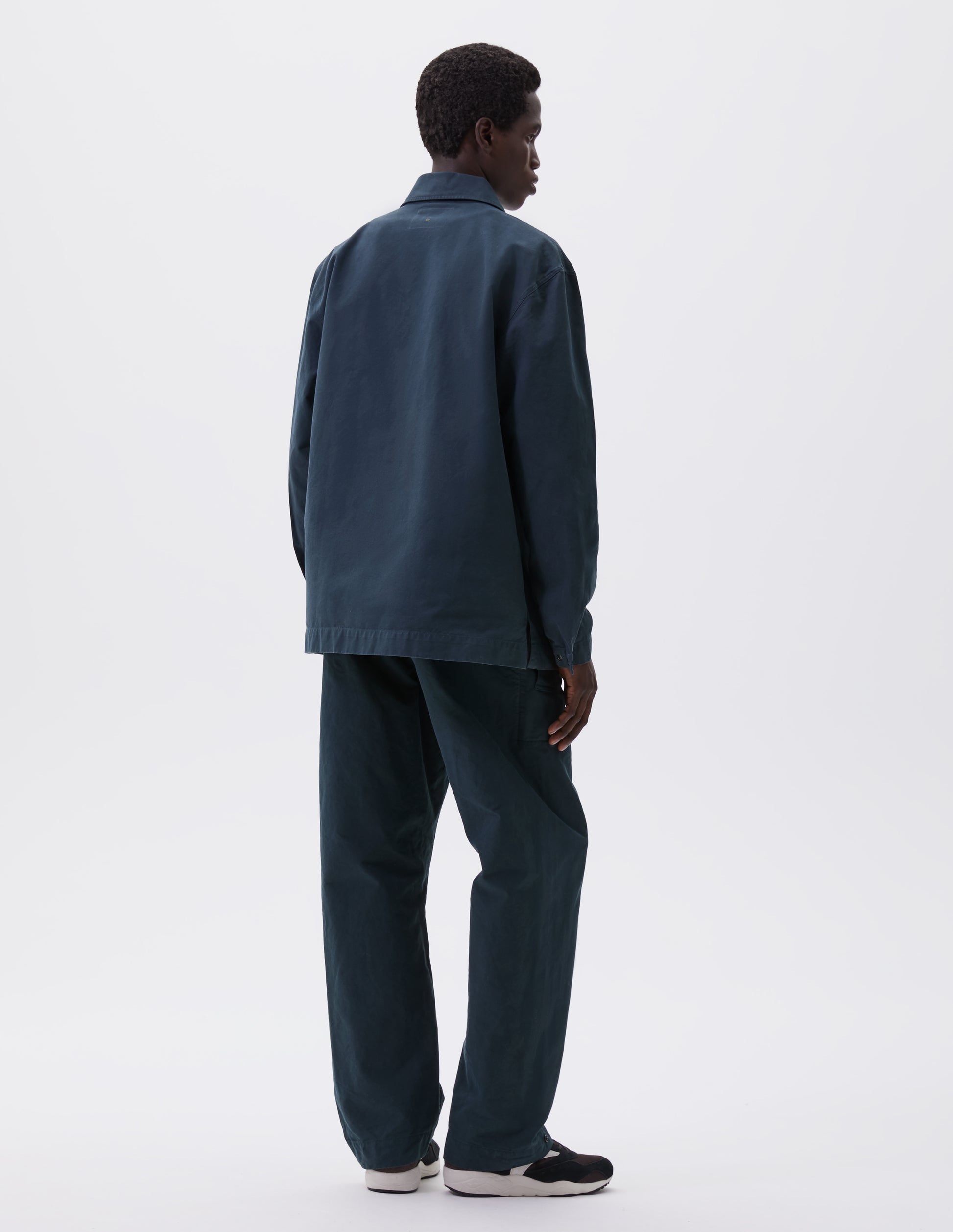 Full body image, back view of model wearing teal blue overshirt with adjustable cuffs and vent split hem. Styled with matching trousers and brown and black trainers.