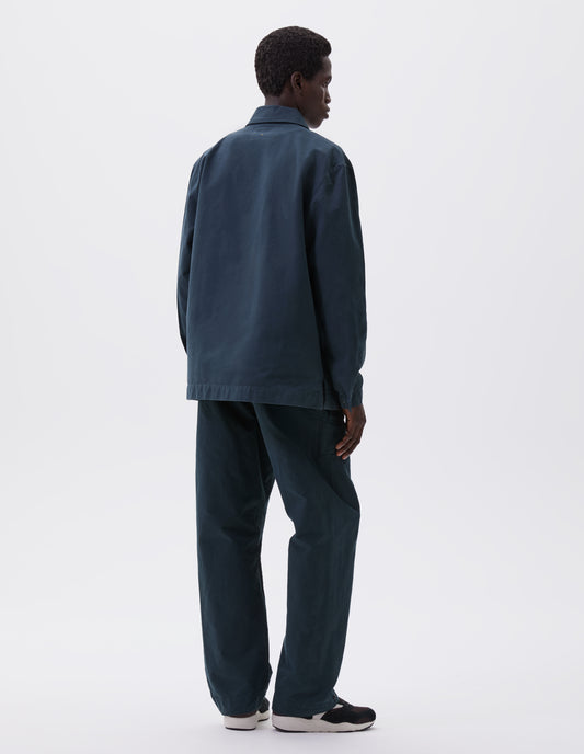 Full body image, back view of model wearing teal blue overshirt with adjustable cuffs and vent split hem. Styled with matching trousers and brown and black trainers.