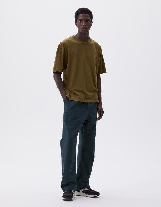 Full body image, front view of model wearing relaxed blue trousers with slant welt pockets, corozo brace button fly and press stud hem adjusters. Styled with green t-shirt and black, brown and white trainers.