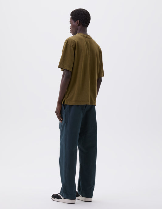 Full body image, back view of model wearing green cotton t-shirt with vent split hem. Styled with blue trousers and brown and black trainers.