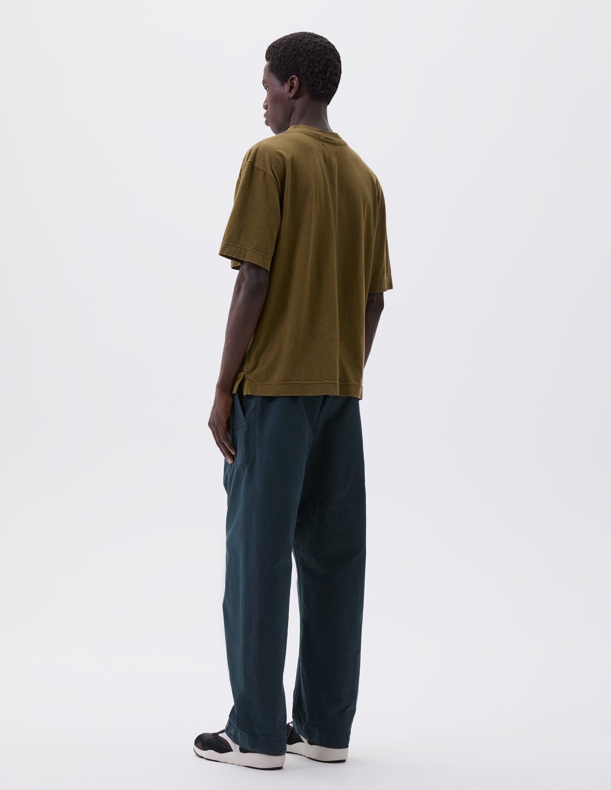 Full body image, back view of model wearing relaxed blue trousers with slant welt pockets  and press stud hem adjusters. Styled with green t-shirt and black, brown and white trainers 