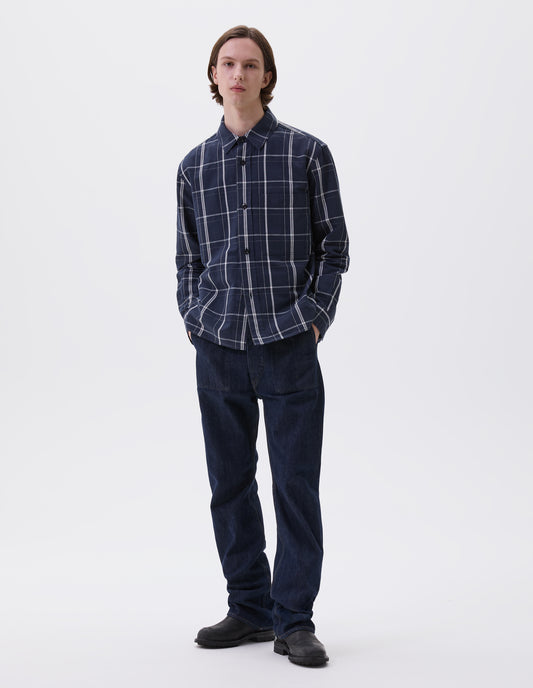 Full body image, front view of model wearing blue and white checked long sleeve shirt. With raised placket, corozo brace buttons and button through chest patch pocket. Styled with indigo blue jeans and black leather boots.