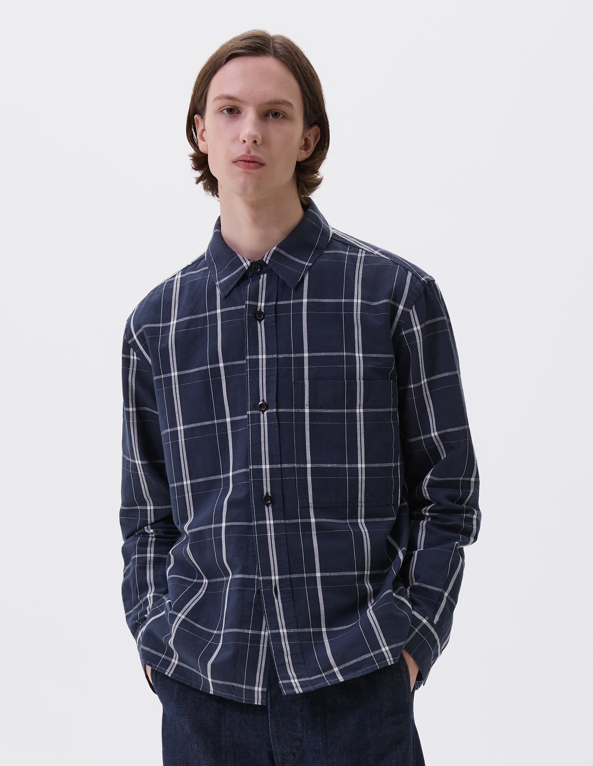 Cropped body image, front view of model wearing blue and white checked long sleeve shirt. With raised placket, corozo brace buttons and button through chest patch pocket. Styled with indigo blue jeans.