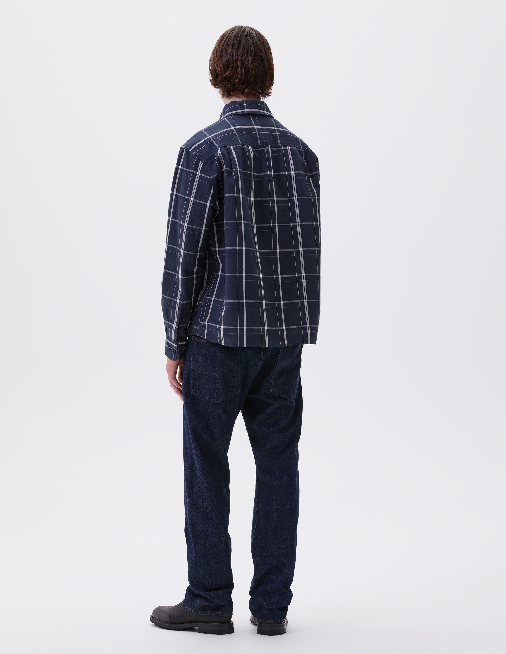 Full body image, back view of model wearing blue and white checked long sleeve shirt. With corozo brace button cuffs and stitch side gusset detailing. Styled with indigo blue jeans and black leather boots.