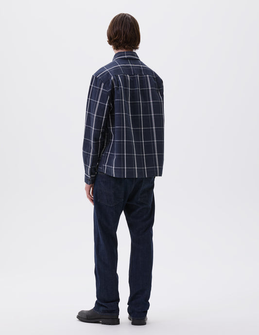 Full body image, back view of model wearing blue and white checked long sleeve shirt. With corozo brace button cuffs and stitch side gusset detailing. Styled with indigo blue jeans and black leather boots.