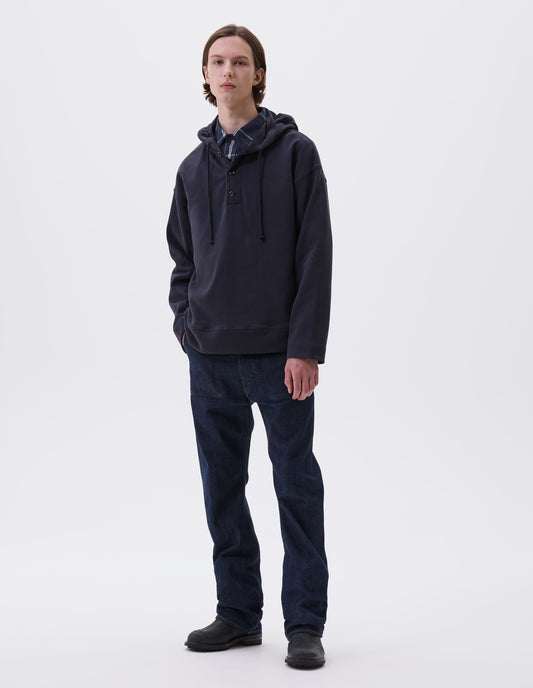 Full body image, front view of model wearing blue black cotton hoodie, with drawcord hood, corozo buttons, straight cuffs and stitch seam detailing. Styled with blue and white checked shirt, indigo blue jeans and black leather boots.