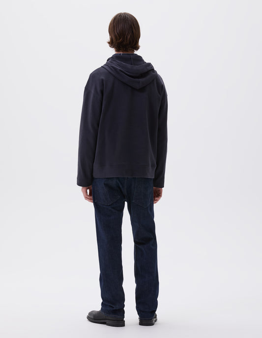 Full body image, back view of model wearing blue black cotton hoodie, with drawcord hood, straight cuffs and stitch seam detailing. Styled with indigo blue jeans and black leather boots.