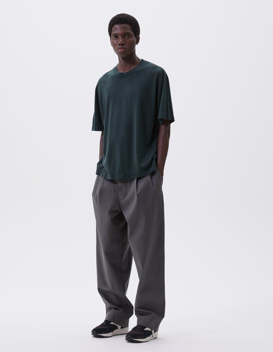 Full body image, front view of model wearing blue green cotton t-shirt with vent split hem. Styled with pleated grey trousers and brown and black trainers. 