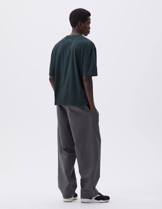 Full body image, back view of model wearing blue green cotton t-shirt with vent split hem. Styled with grey trousers and brown and black trainers. 