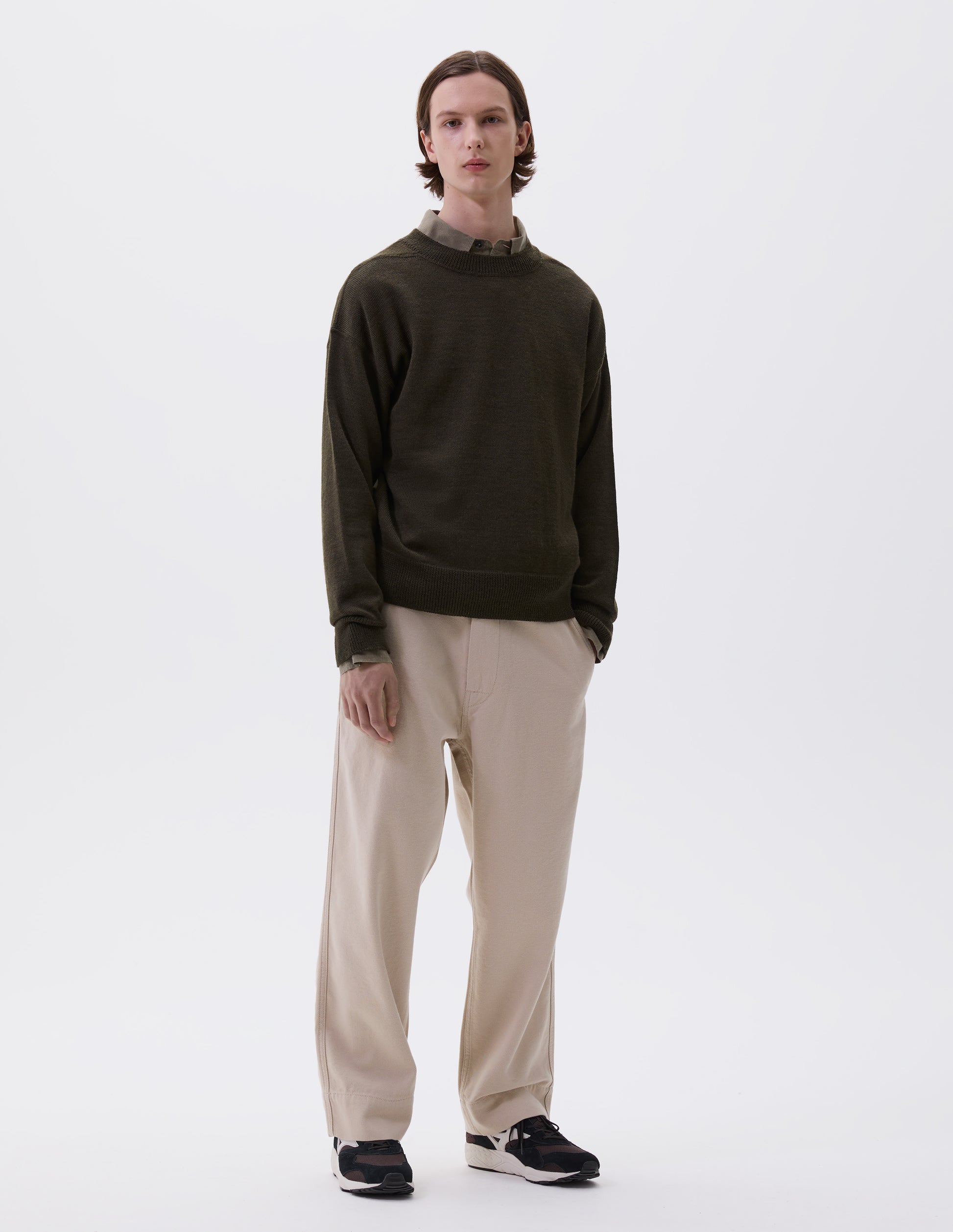 Full body image, front view of model wearing green wool jumper, with contrasting rib on neck, cuffs and hem and flatlock stitching detailing on seams. Styled over green shirt, beige trousers and brown and black trainers. 