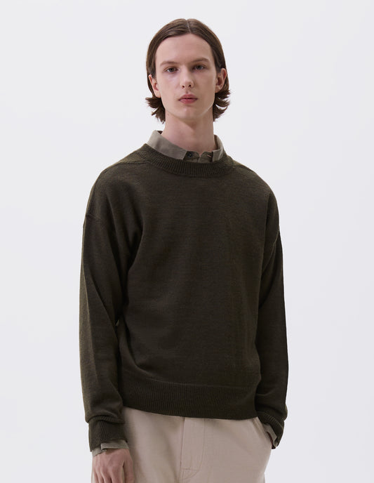 Cropped body image, front view of model wearing green wool jumper, with contrasting rib on neck, cuffs and hem and flatlock stitching detailing on seams. Styled over green shirt and beige trousers.