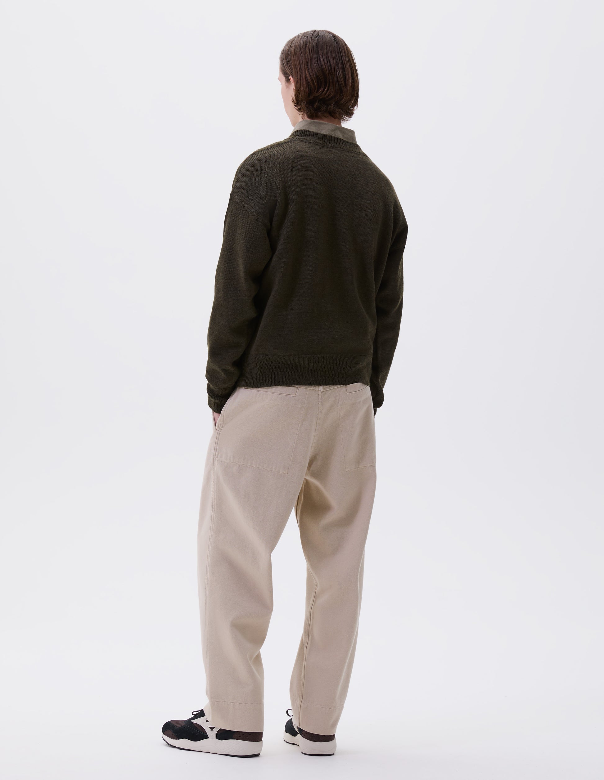 Full body image, back view of model wearing green wool jumper, with contrasting rib on neck, cuffs and hem, flatlock stitching detailing on seams and t shape shoulder seams. Styled over green shirt, beige trousers and brown and black trainers. 