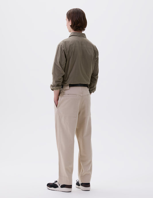 Full body image, back view of model wearing beige trousers  with side elasticated waistband, wide belt loops and stitch through welt back pockets on back yoke. Styled with green shirt, black leather belt and black and brown trainers. 