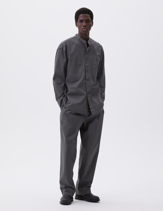 Full body image, front view of model wearing dark grey cotton collarless shirt, with stitch through placket, button-through patch pocket and curved side gusset. Styled with grey trousers and black leather boots.