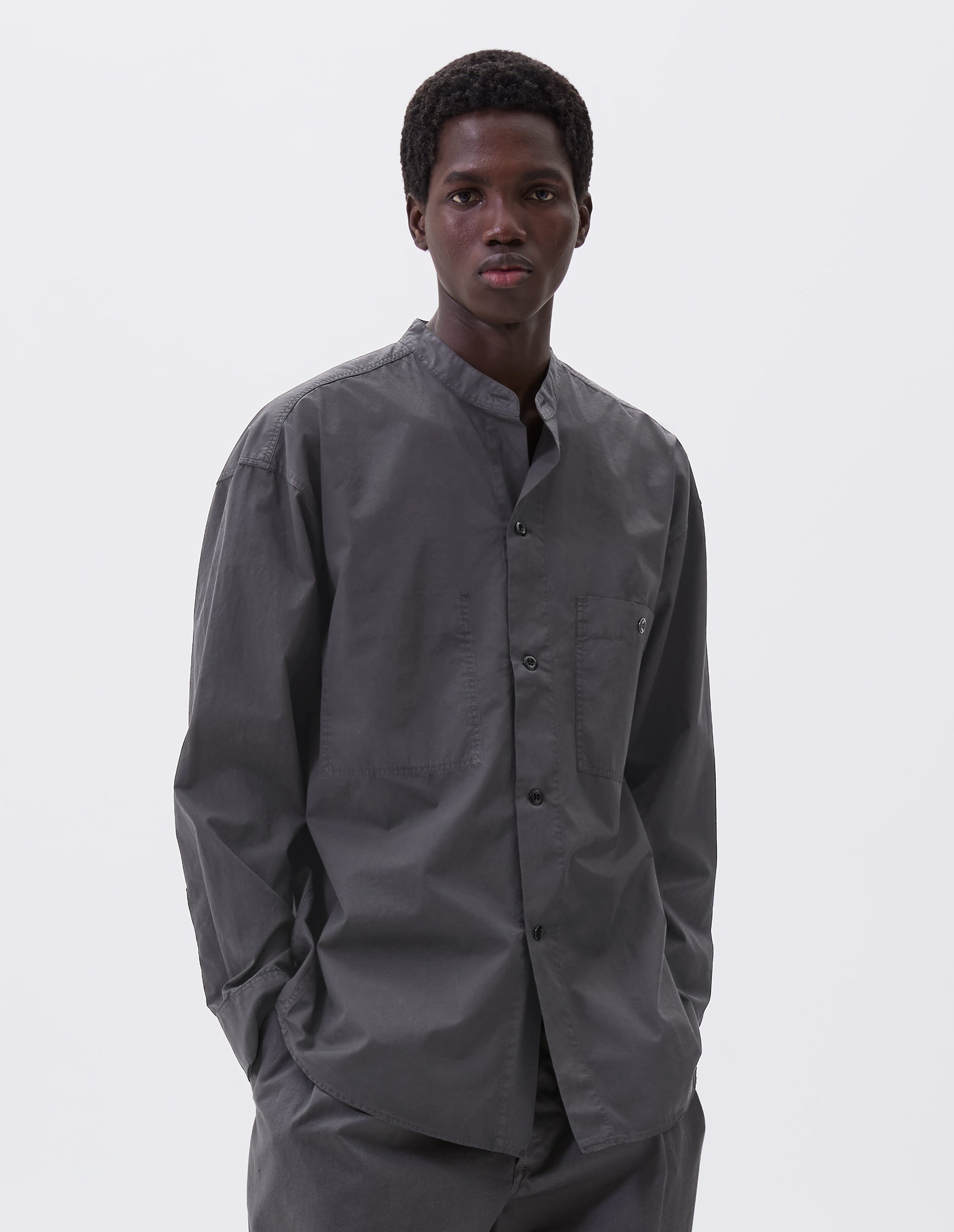 Cropped body image, front view of model wearing dark grey cotton collarless shirt, with stitch through placket, button-through patch pocket and curved side gusset. Styled with grey trousers.