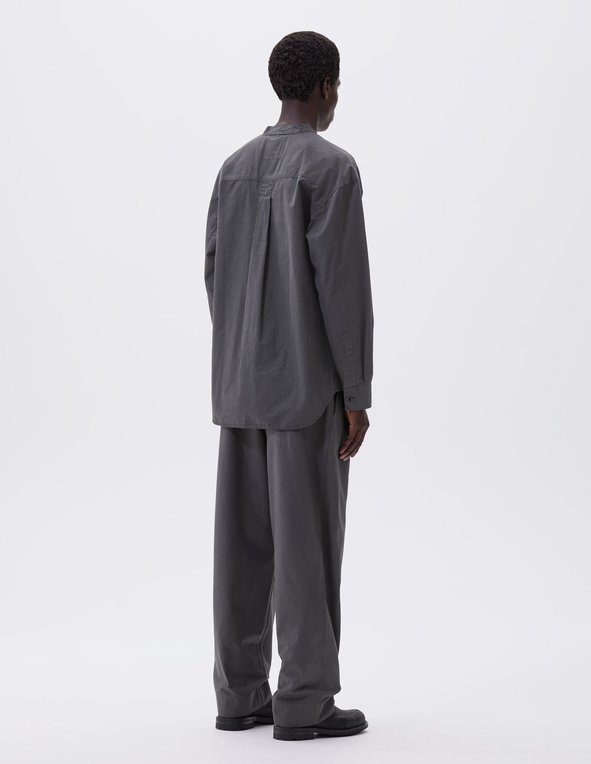 Full body image, back view of model wearing dark grey cotton collarless shirt, with curved side gusset, box pleat on back yoke and corozo brace button cuffs. Styled with grey trousers and black leather boots.