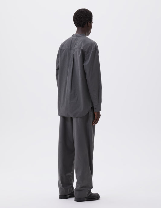 Full body image, back view of model wearing dark grey cotton collarless shirt, with curved side gusset, box pleat on back yoke and corozo brace button cuffs. Styled with grey trousers and black leather boots.