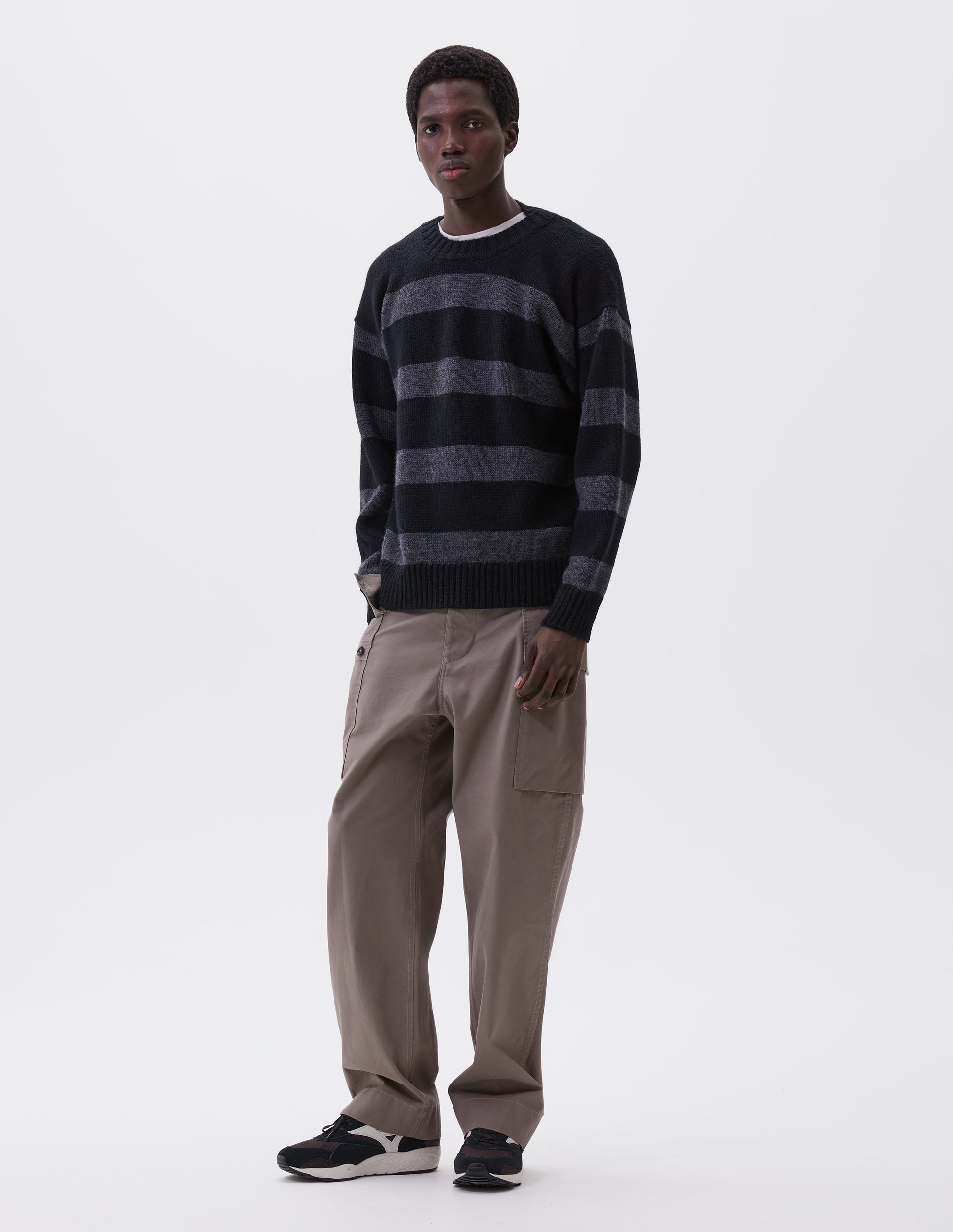 Full body image, front view of model wearing black and grey striped wool jumper, with ribbed neck, cuffs and hem. Styled over white t-shirt, with beige trousers and black and brown trainers.