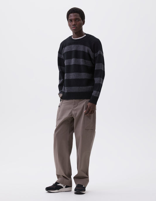 Full body image, front view of model wearing black and grey striped wool jumper, with ribbed neck, cuffs and hem. Styled over white t-shirt, with beige trousers and black and brown trainers.