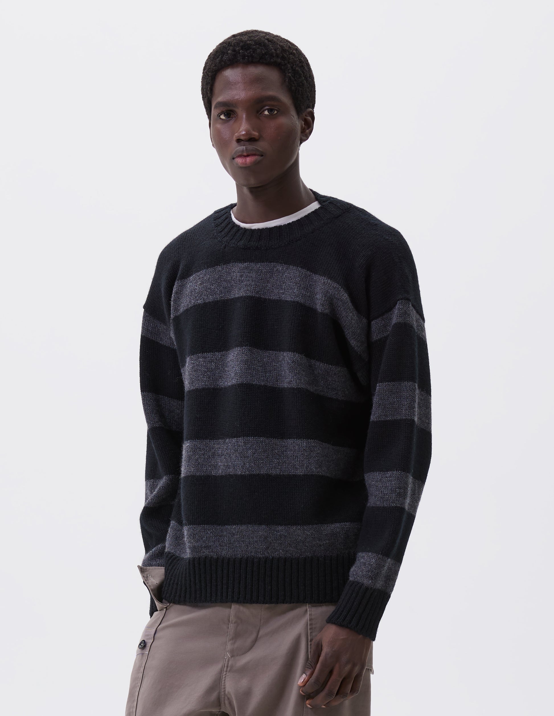 Cropped body image, front view of model wearing black and grey striped wool jumper, with ribbed neck, cuffs and hem. Styled over white t-shirt, with beige trousers.