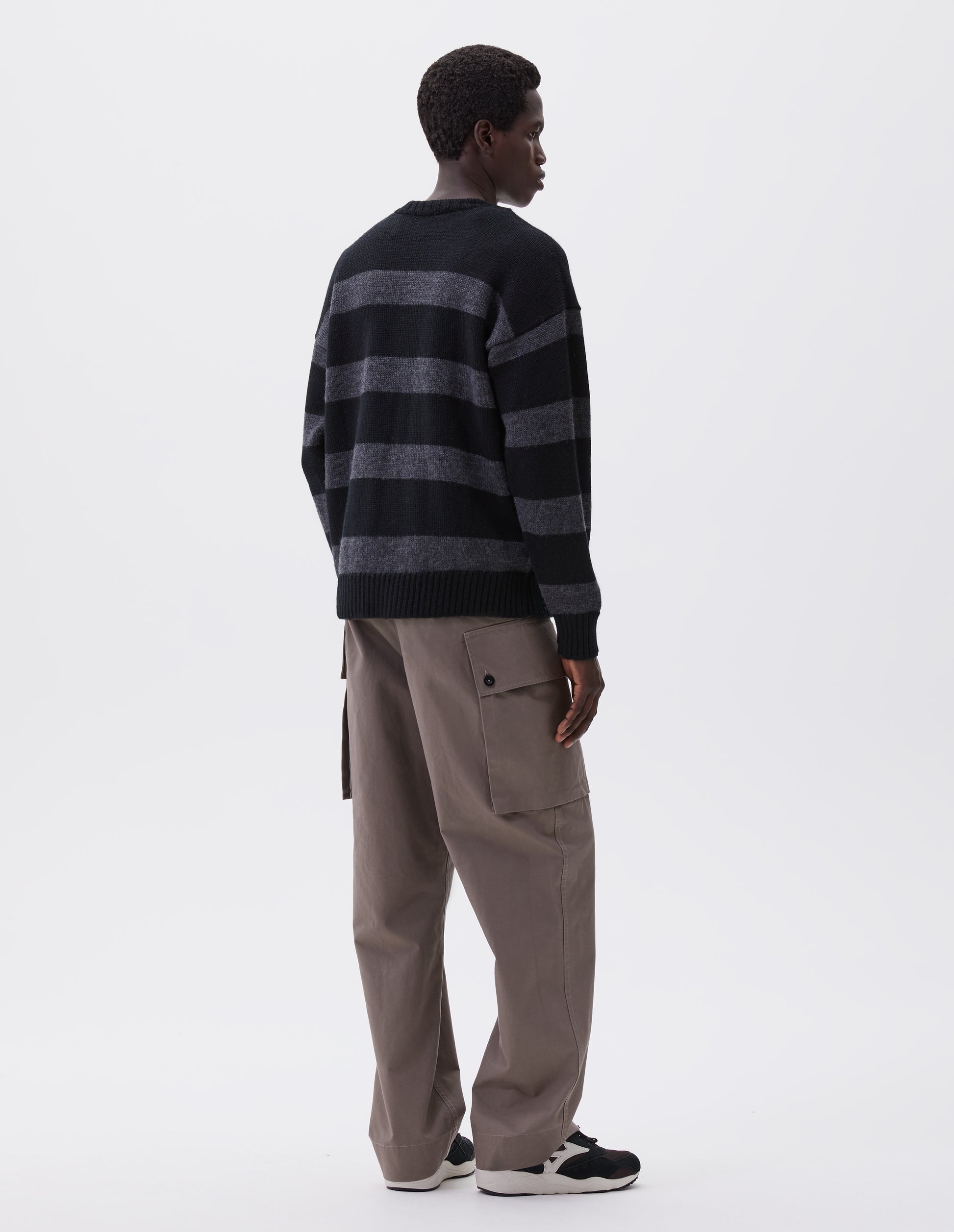 Full body image, back view of model wearing black and grey striped wool jumper, with ribbed neck, cuffs and hem. Styled with beige trousers and black and brown trainers.