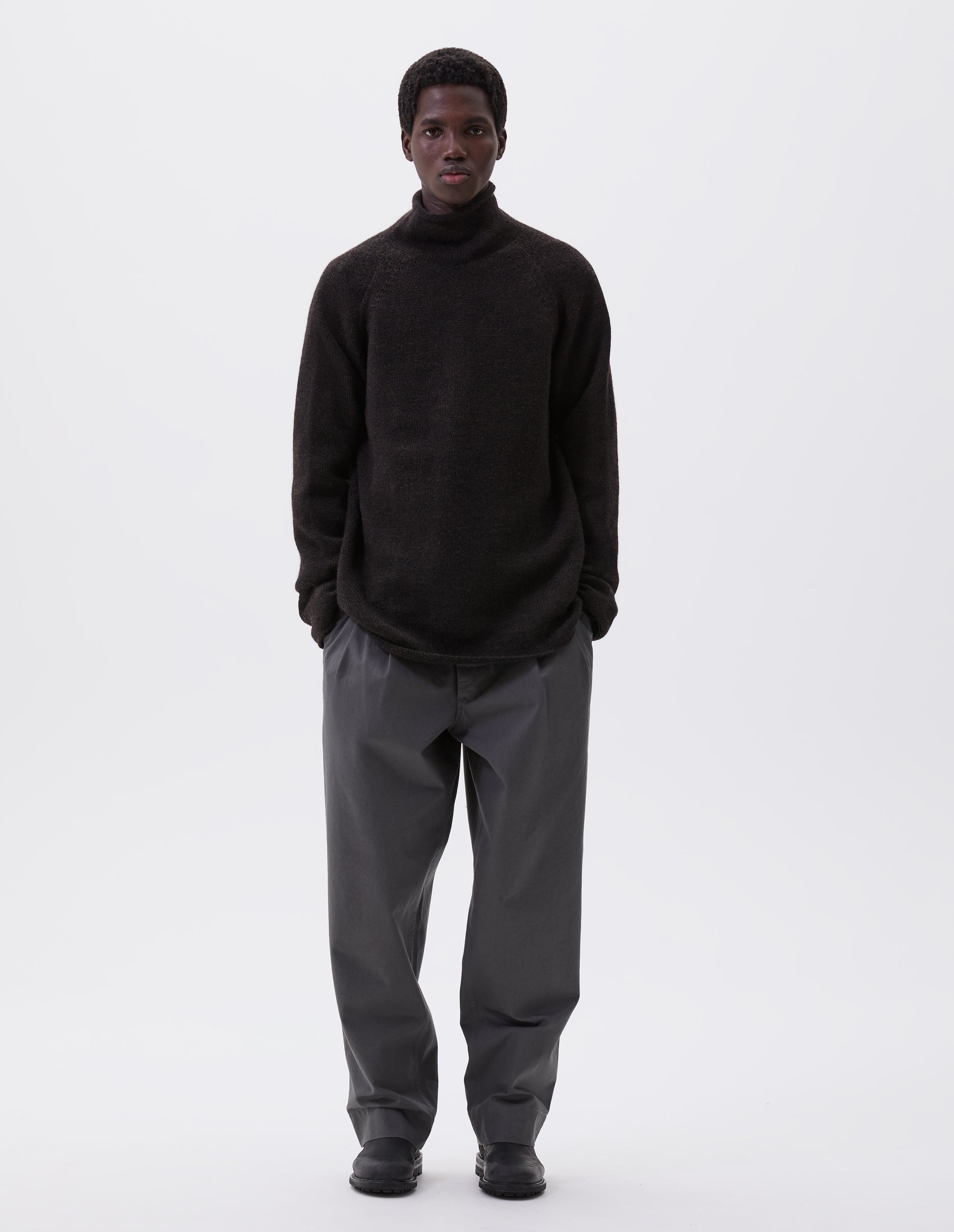 Full body image, front view of model wearing brown roll neck jumper with seamless construction, high neck, raglan sleeve and straight cuffs and hem. Styled with grey trousers and black leather boots.