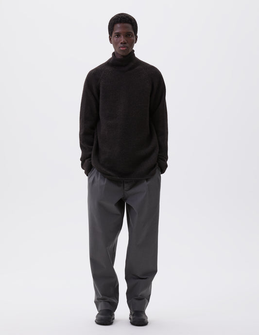 Full body image, front view of model wearing brown roll neck jumper with seamless construction, high neck, raglan sleeve and straight cuffs and hem. Styled with grey trousers and black leather boots.