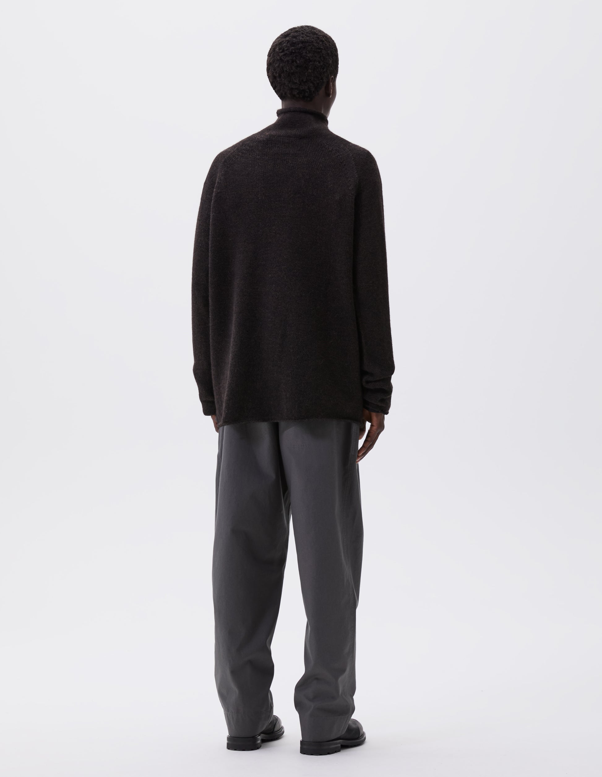 Full body image, back view of model wearing brown roll neck jumper with seamless construction, high neck, raglan sleeve and straight cuffs and hem. Styled with grey trousers and black leather boots.