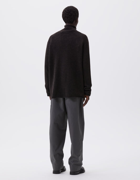 Full body image, back view of model wearing brown roll neck jumper with seamless construction, high neck, raglan sleeve and straight cuffs and hem. Styled with grey trousers and black leather boots.