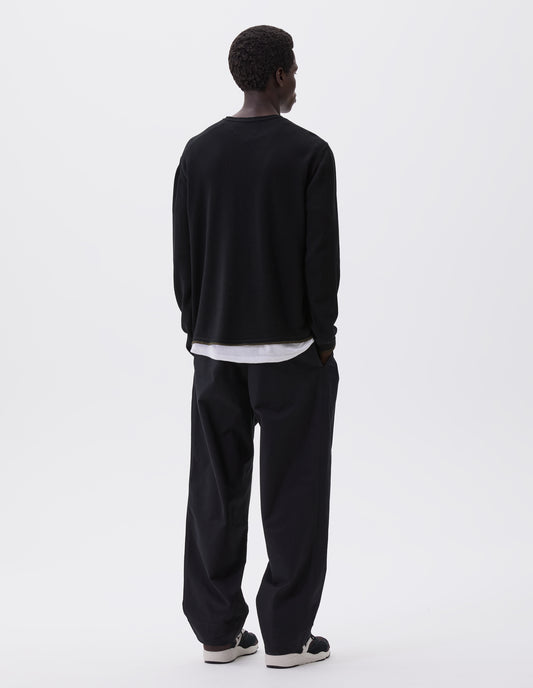 Full body image, back view of model wearing black long sleeve knitted thermal with ribbed collar, khaki green stripe on cuffs and hem, styled with thermal over white t-shirt, black trousers and black, brown and white trainers. 