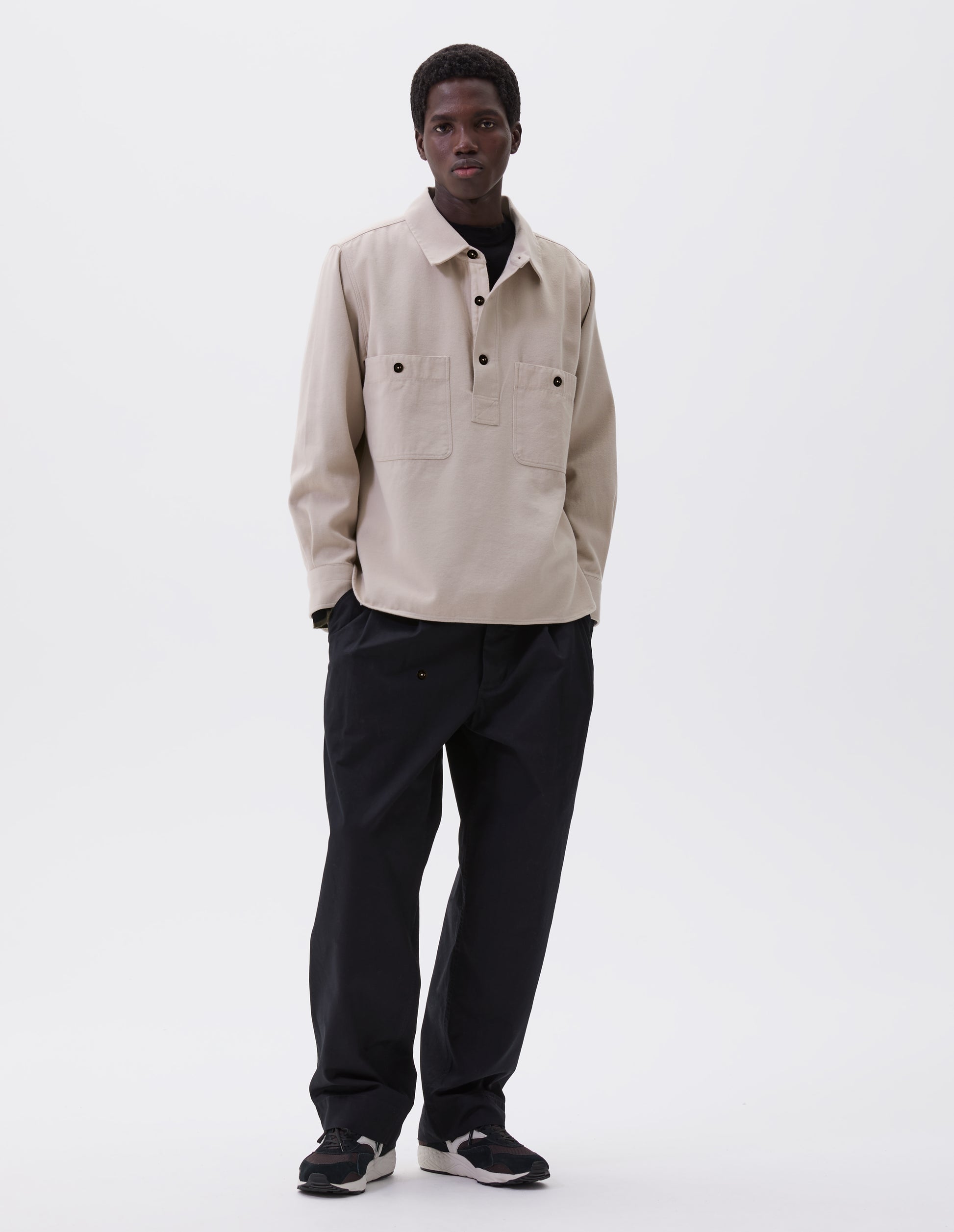 Full body image, front view of model wearing beige overhead shirt with raised placket, box stitch detailing, button through chest patch pockets and elongated back hem. Styled over black mock neck top, black trousers and brown and black trainers.