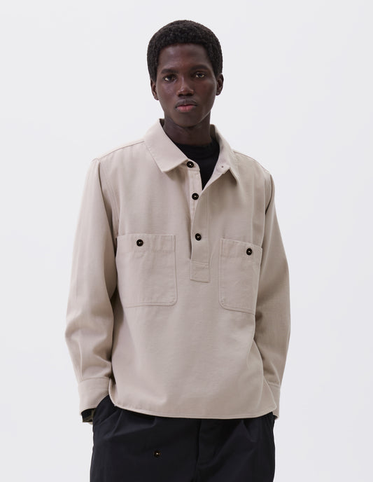 Cropped body image, front view of model wearing beige overhead shirt with raised placket, box stitch detailing, button through chest patch pockets and elongated back hem. Styled over black mock neck top and black trousers.
