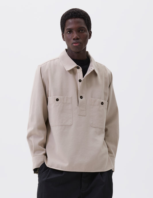 Cropped body image, front view of model wearing beige overhead shirt with raised placket, box stitch detailing, button through chest patch pockets and elongated back hem. Styled over black mock neck top and black trousers.