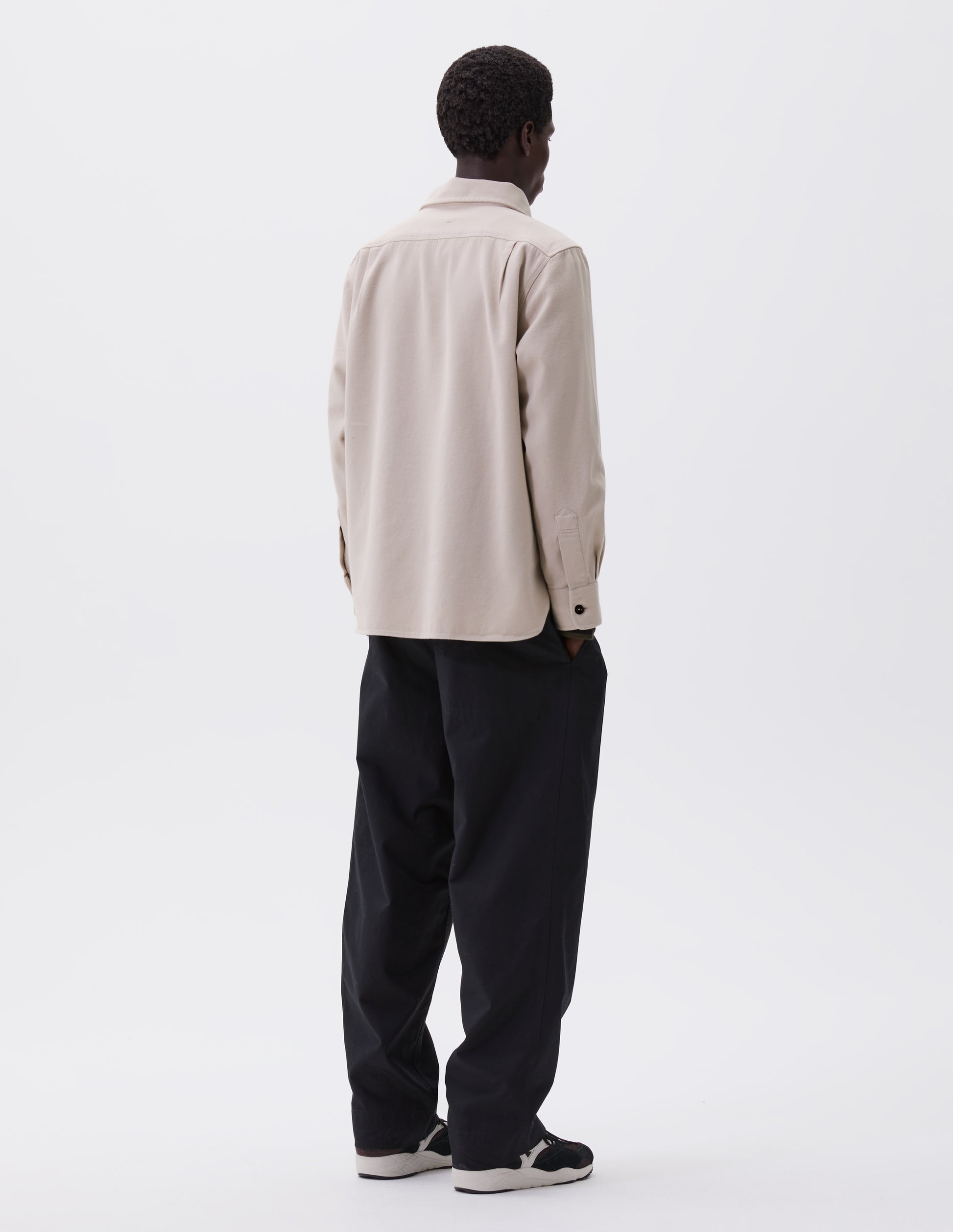 Full body image, back view of model wearing beige overhead shirt with raised placket, box stitch detailing, button through chest patch pockets and elongated back hem. Styled with black trousers and brown and black trainers.