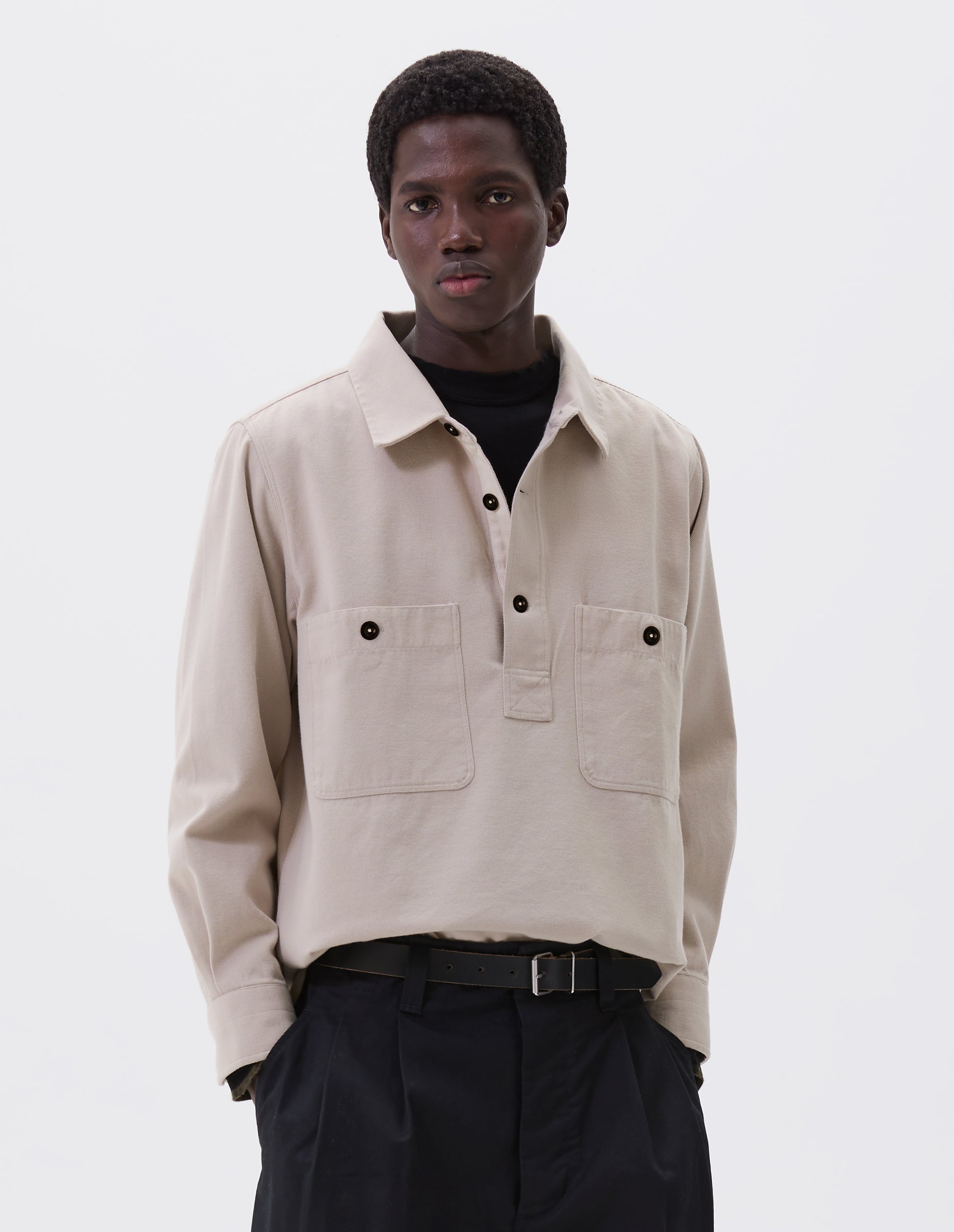 Cropped body image of male model wearing black leather belt with simple silver roller buckle and metal keeper. Styled with black t-shirt, beige overshirt and black pleated trousers.