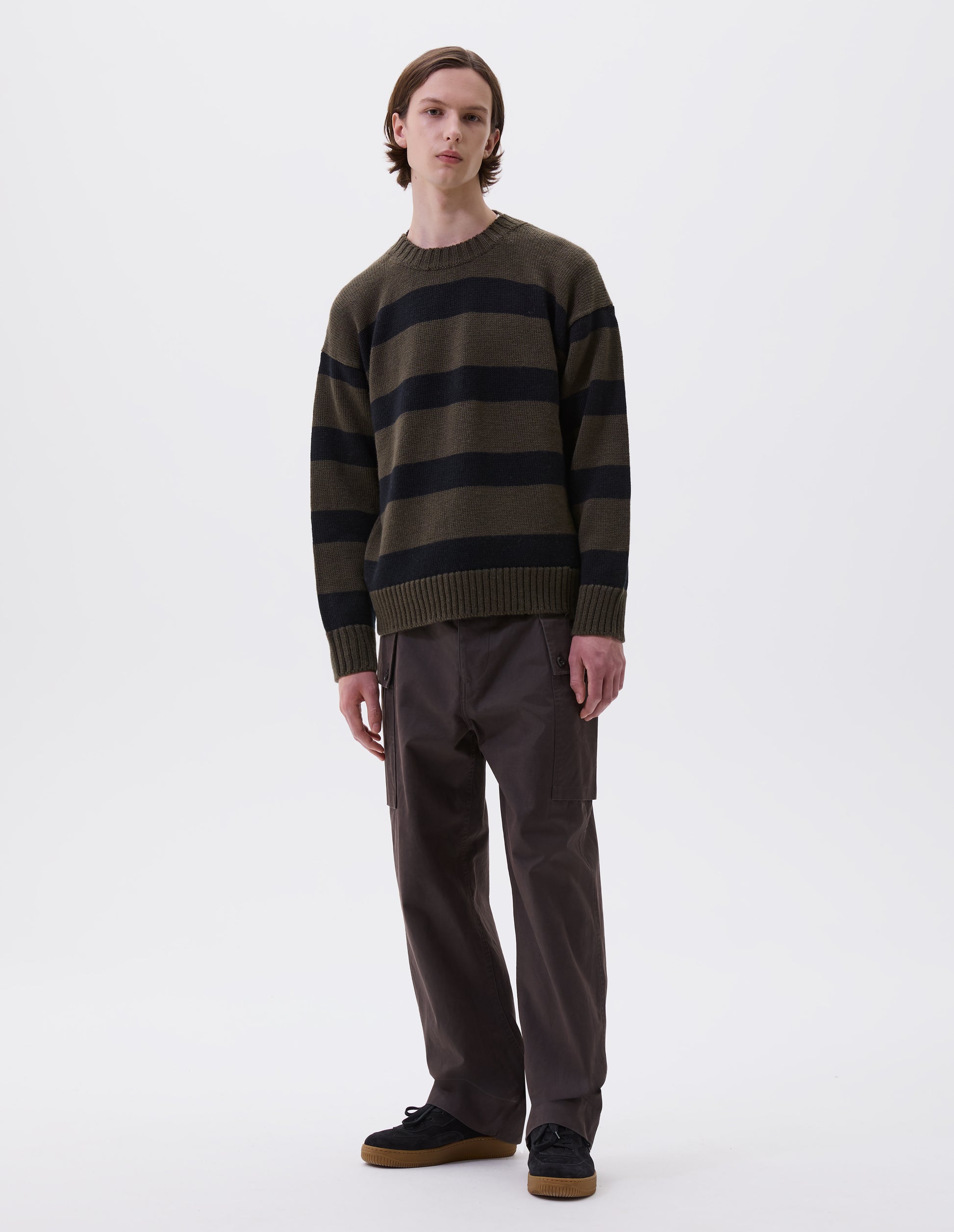 Full body image, front view of model wearing green and black striped wool jumper, with ribbed neck, cuffs and hem. Styled with brown trousers and black suede trainers.