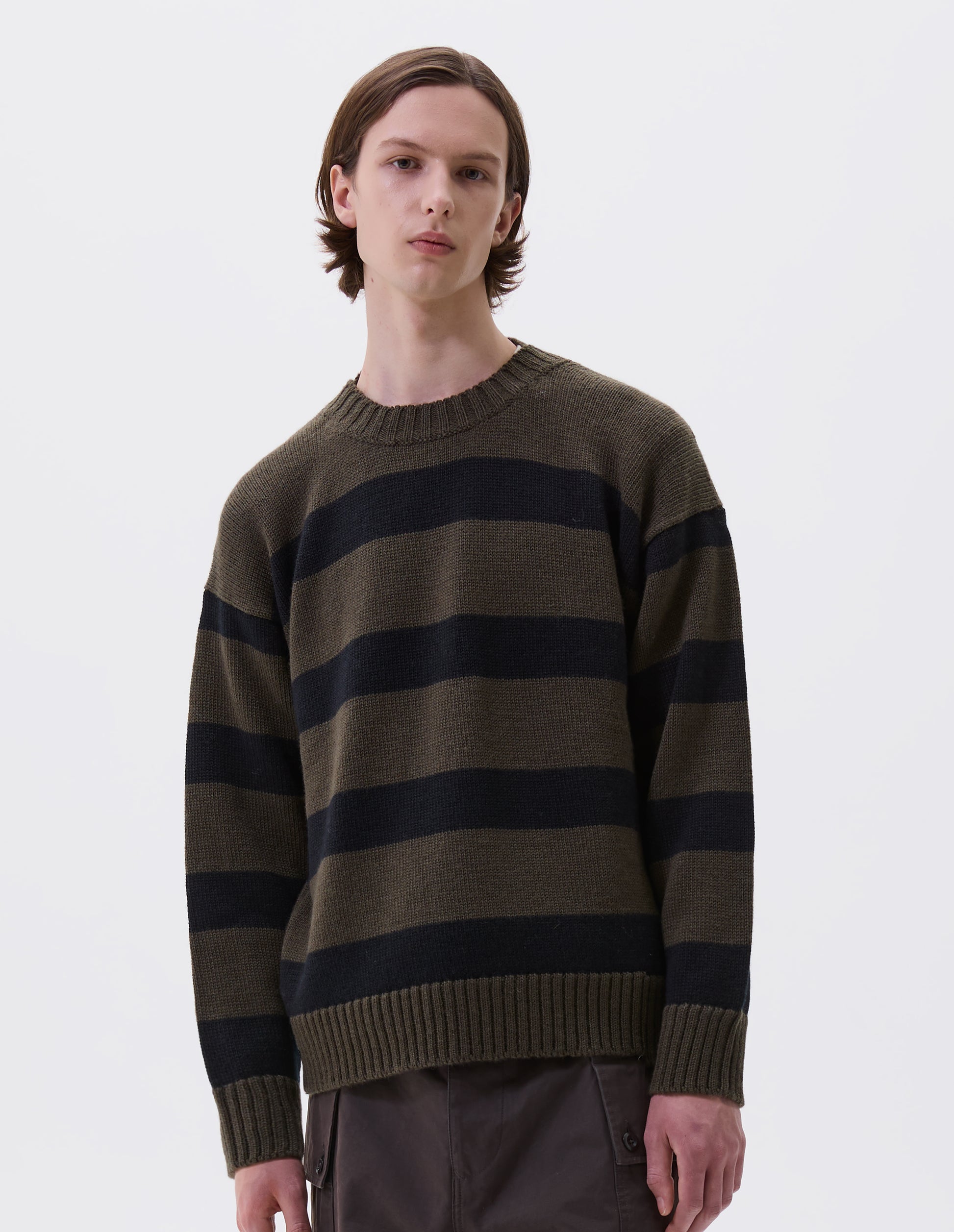 Cropped body image, front view of model wearing green and black striped wool jumper, with ribbed neck, cuffs and hem. Styled with brown trousers.