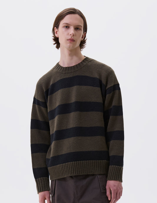 Cropped body image, front view of model wearing green and black striped wool jumper, with ribbed neck, cuffs and hem. Styled with brown trousers.