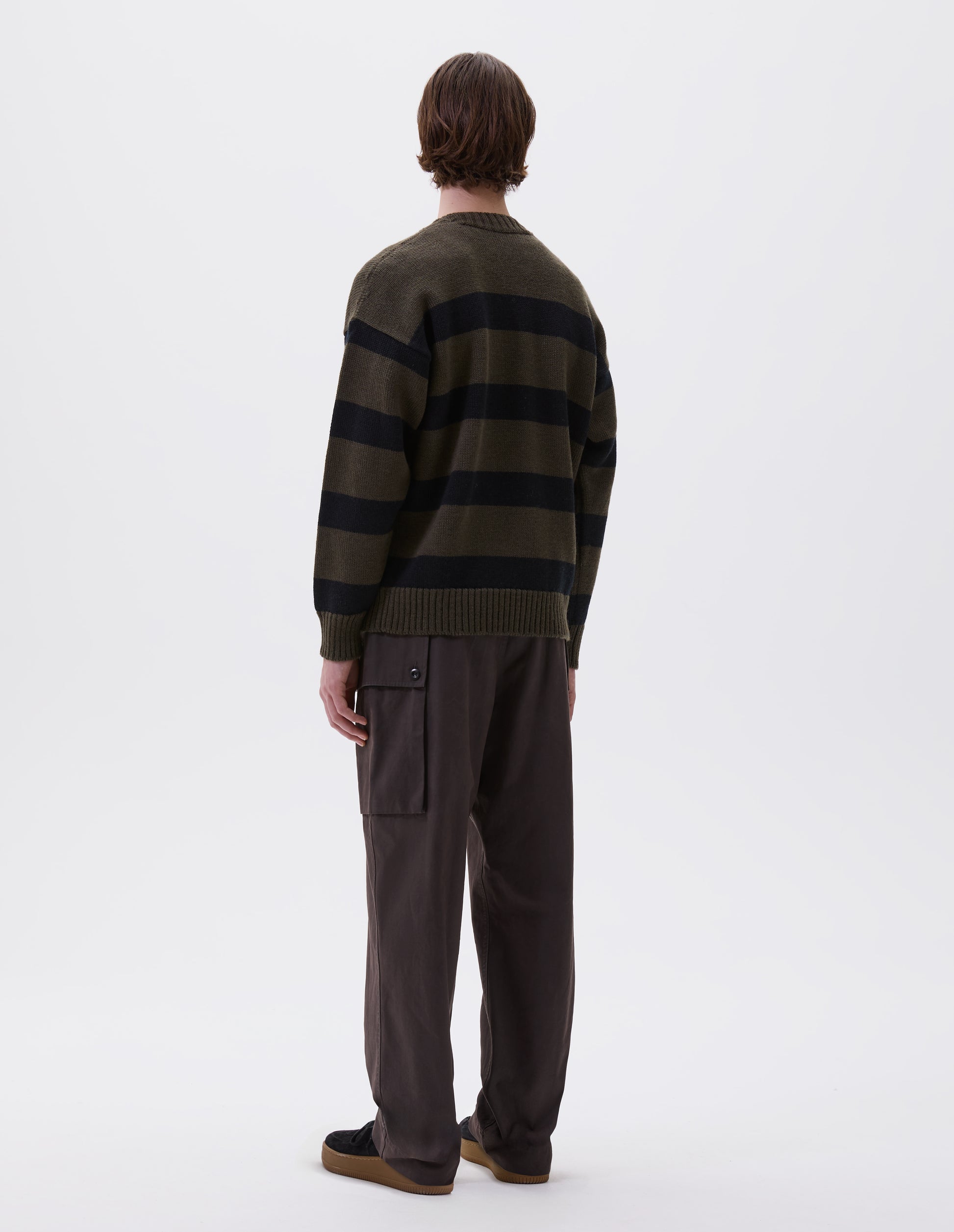 Full body image, back view of model wearing green and black striped wool jumper, with ribbed neck, cuffs and hem. Styled with brown trousers and black suede trainers.