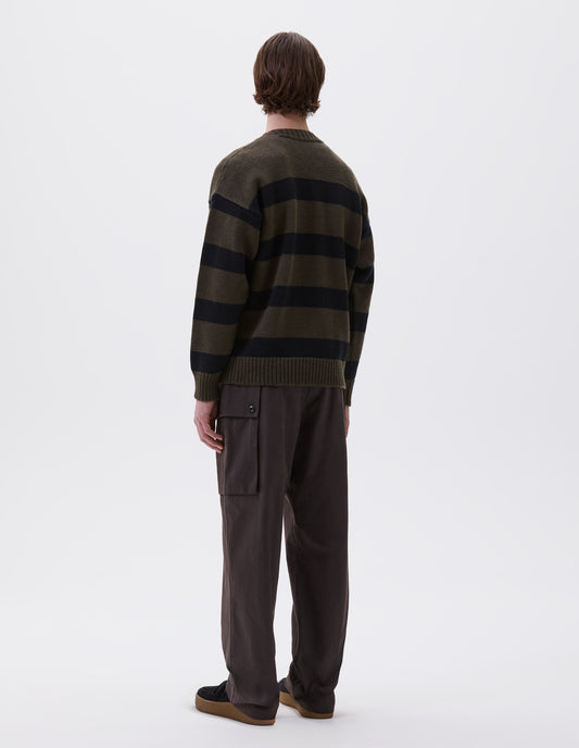 Full body image, back view of model wearing green and black striped wool jumper, with ribbed neck, cuffs and hem. Styled with brown trousers and black suede trainers.