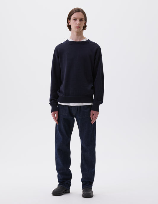 Full body image, front view of model wearing dark navy wool jumper, with contrasting rib on neck, cuffs and hem and flatlock stitching detailing on seams. Styled over white t-shirt, indigo denim jeans and black leather boots.