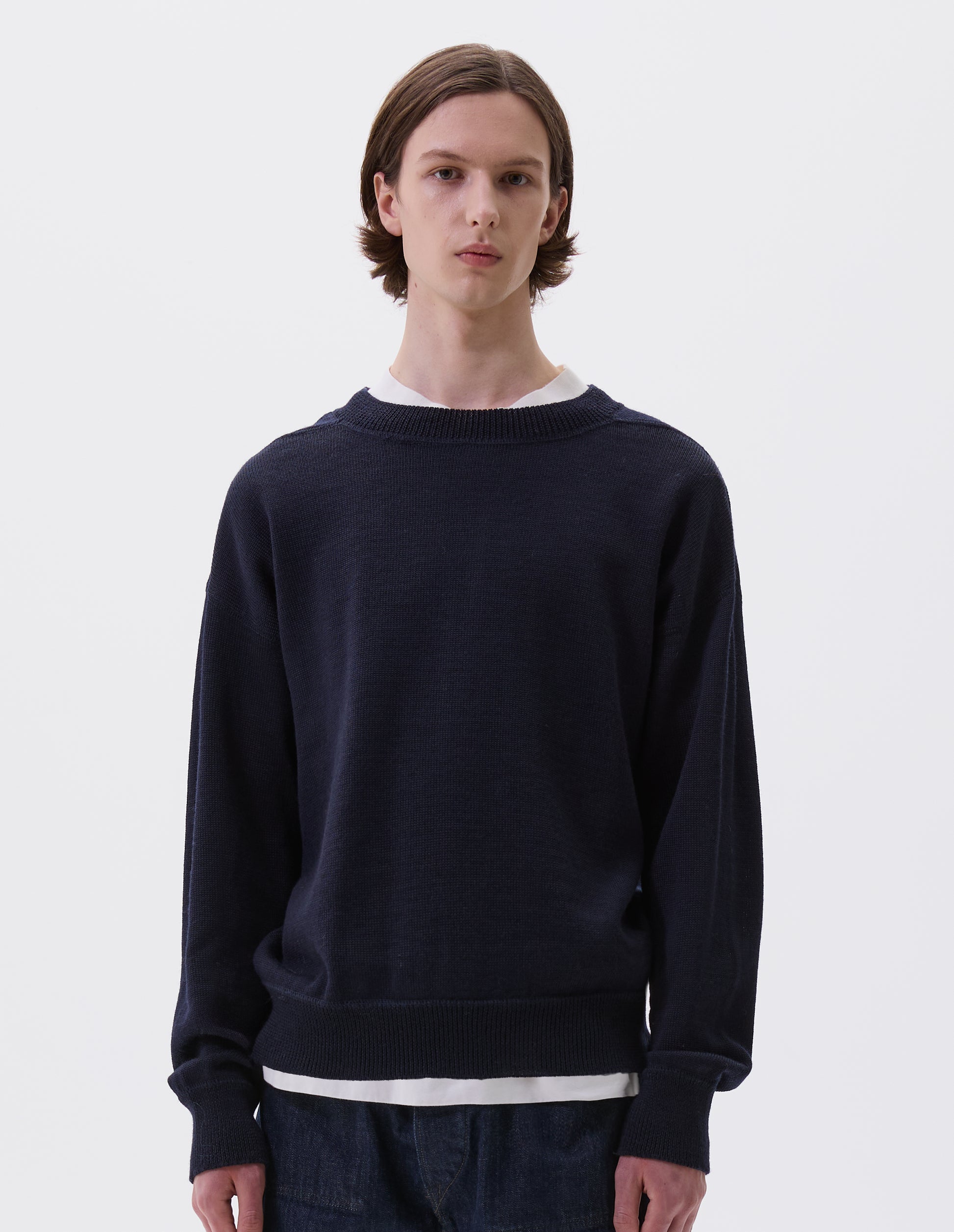 Cropped body image, front view of model wearing dark navy wool jumper, with contrasting rib on neck, cuffs and hem and flatlock stitching detailing on seams. Styled over white t-shirt and indigo denim jeans.