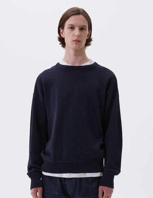 Cropped body image, front view of model wearing dark navy wool jumper, with contrasting rib on neck, cuffs and hem and flatlock stitching detailing on seams. Styled over white t-shirt and indigo denim jeans.