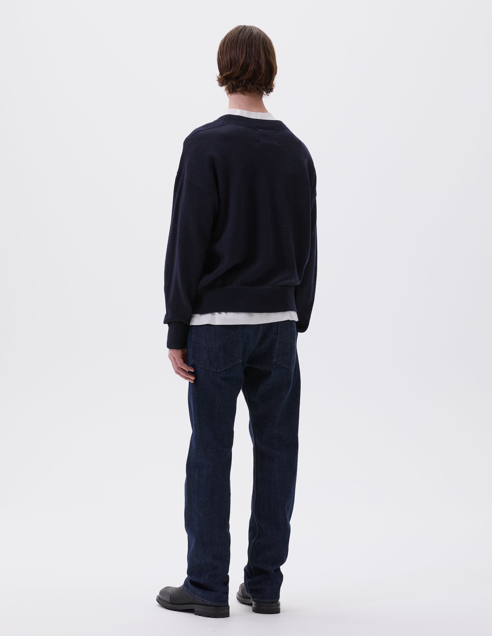Full body image, back view of model wearing dark navy wool jumper, with contrasting rib on neck, cuffs and hem, flatlock stitching detailing on seams and t shape shoulder seams. Styled over white t-shirt, indigo denim jeans and black leather boots.