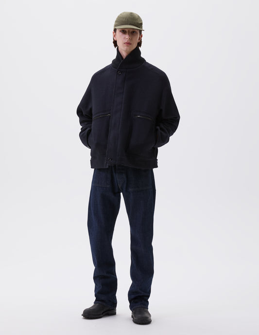 Full body image, front view of model wearing indigo denim twill jeans, with metal button fly, grown on waistband with belt loops and stitch-through slant pockets. Styled with green cap, navy blue jacket and black leather boots.