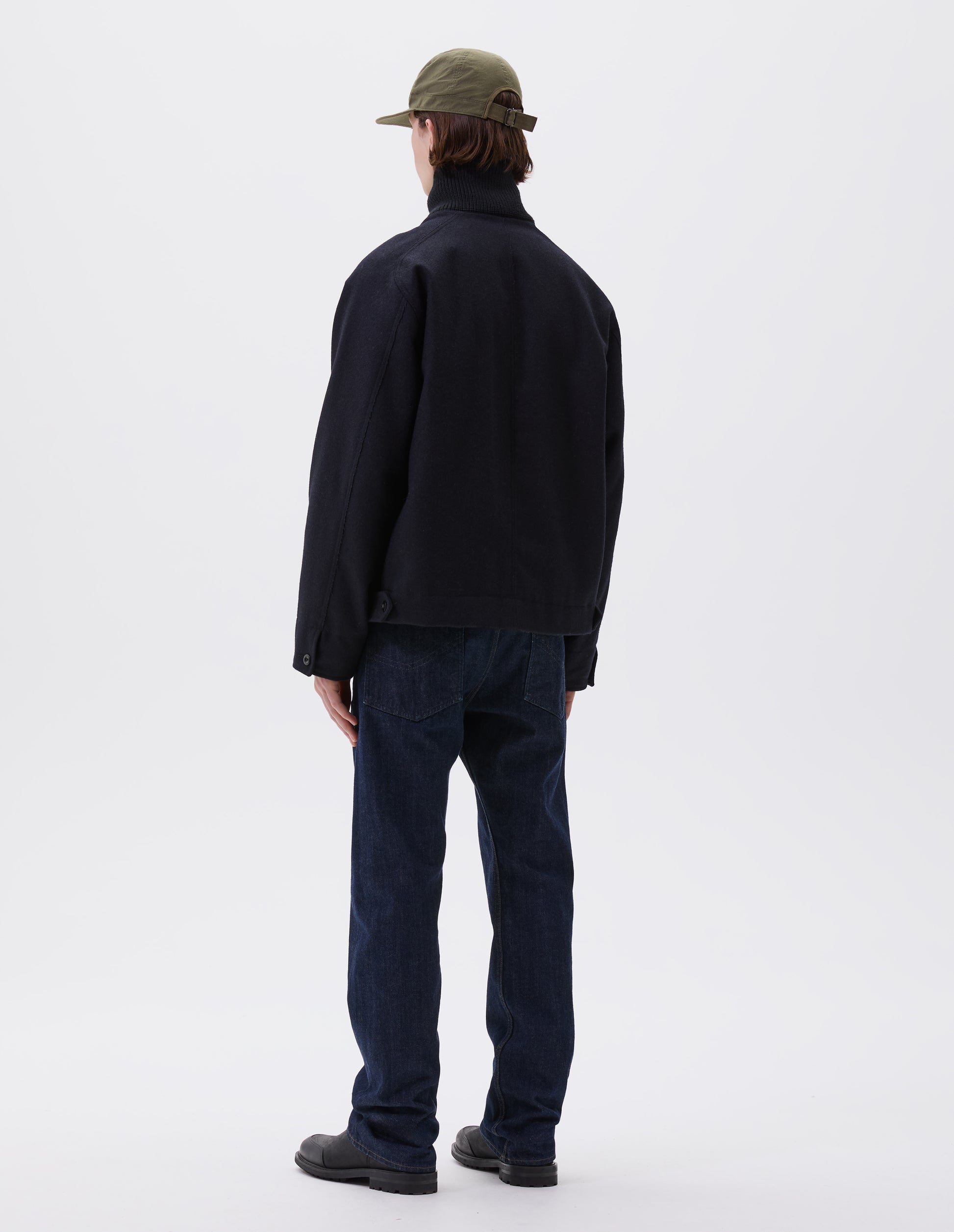 Full body image, back view of model wearing navy blue rib neck jacket, with contrast wool rib collar, saddle sleeves, button through cuffs, raw edge detailing and button tabs to adjust the hem. Styled with indigo blue jeans, green cotton cap and black leather boots.