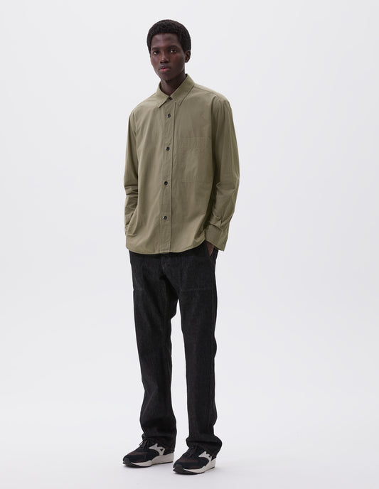 Full body image, front view of model wearing green brushed cotton shirt with raised placket, large chest pocket, corozo button closure and side gusset stitch detailing. Styled with black denim jeans and black and brown trainers.
